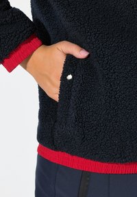 Veste en polaire marine avec des poignets et un ourlet élastiques rouges. Dispose d'une poche latérale avec un détail de bouton blanc visible. Matériau doux et texturé.