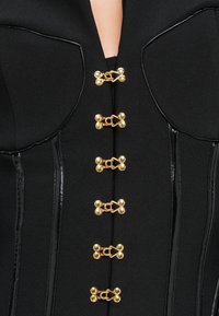 Μαύρο δομημένο ρούχο με γυαλιστερό μαύρο περικόχλιο και έξι συμμετρικούς χρυσούς κρίκους κλειδώματος κατά μήκος του κέντρου μπροστά.