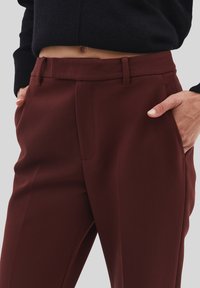 Primo piano di una persona che indossa pantaloni bordeaux a vita alta con le mani in tasca e un maglione corto nero che mostra un ventre scoperto.