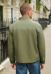 Olive green, lightweight jacket with a relaxed fit, button cuffs, and a back yoke. Worn over blue jeans, shown from the back.