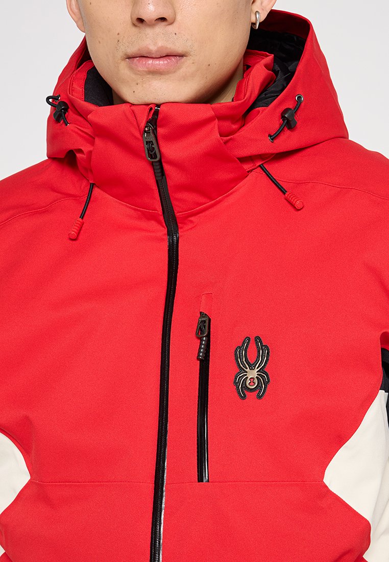 Rote wasserdichte Jacke mit Reißverschluss, verstellbare Kapuze, schwarzen Akzenten und einem Spinnen-Logo. Glatte Textur mit sportlichem Design.