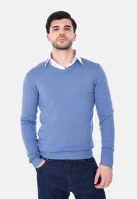 Giovane uomo con capelli e barba scuri che indossa un maglione blu a scollo a V sopra una camicia bianca con colletto, in piedi con una mano in tasca su uno sfondo neutro.