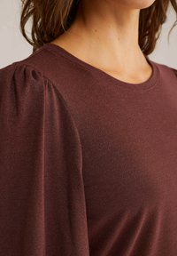 Ženska oblečena v rebrasto maroon bluzo s napihnjenimi rameni in okroglo ovratnikom, ki razkriva vrat in del obraza.