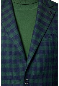 Zielono-granatowa marynarka w kratę z wykrojem na klapy, z pojedynczym czarnym guzikiem i gładkim zielonym swetrem pod spodem.