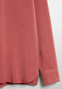 Lys pink skjorte med lange ærmer, en rundet kant, og diskret logo broderi på brystet. Materialet ser blødt ud med en glat struktur.