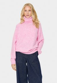 Rožinė tekstūrinė megztinė su aukštu apykakliniu užsegimu ir laisvu siluetu, derinama su tamsiai mėlynomis plačiomis kelnių. Paprastas, modernus dizainas.