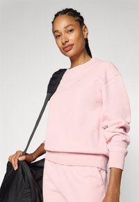 Sweatshirt côtelé rose clair avec des manches et un ourlet. Le modèle tient un sac noir, doté d'une sangle texturée. Tissu lisse, design à coupe décontractée.