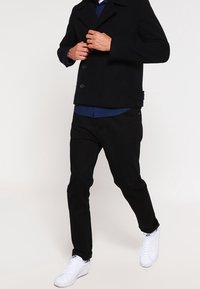 Schwarzer Peacoat mit doppelt geknöpften Knöpfen, kombiniert mit dunkler Hose und weißen Sneakern. Darunter ein blaues Hemd mit Kragen.