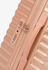 Plano cerrado de una maleta rígida color melocotón con textura acanalada, cierre dorado, cerradura de combinación integrada y asa lateral.