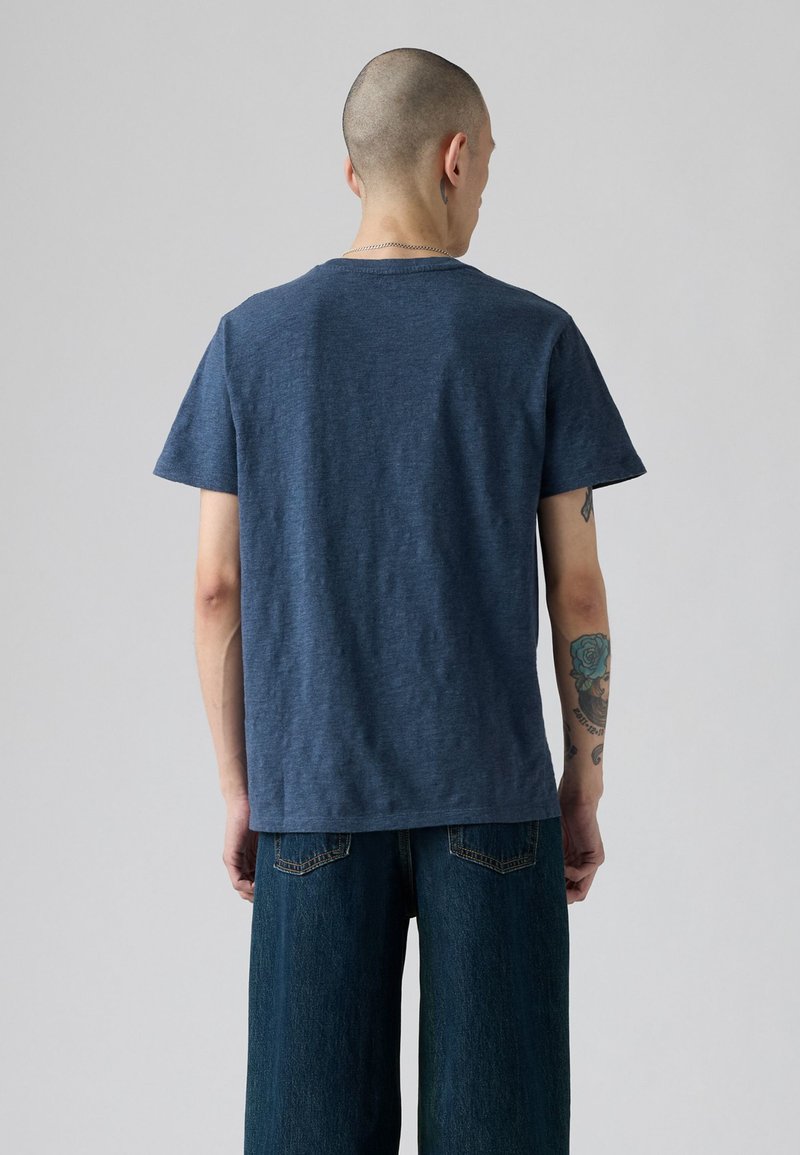 Mann mit kahlem Kopf, der ein schlichtes blaues T-Shirt und dunkelblaue Jeans trägt, Rückansicht vor einem hellgrauen Hintergrund.