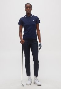 Γυναίκα που στέκεται με μπαστούνι γκολφ, φορώντας μπλε πουκάμισο πόλο, μπλε παντελόνια, λευκό γάντι γκολφ και λευκά παπούτσια γκολφ, με φόντο απλό.