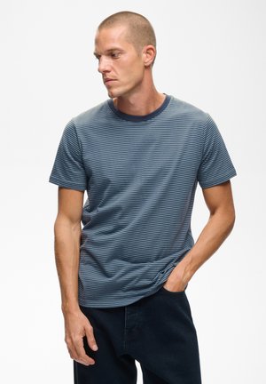 Homme aux cheveux courts portant un t-shirt rayé bleu et gris et un pantalon foncé, regardant vers sa gauche avec une main dans sa poche.