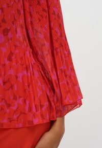 Blouse rouge transparente avec tissu plissé et motif abstrait rose. Les manches sont fluides, légèrement translucides, avec un bord superposé.