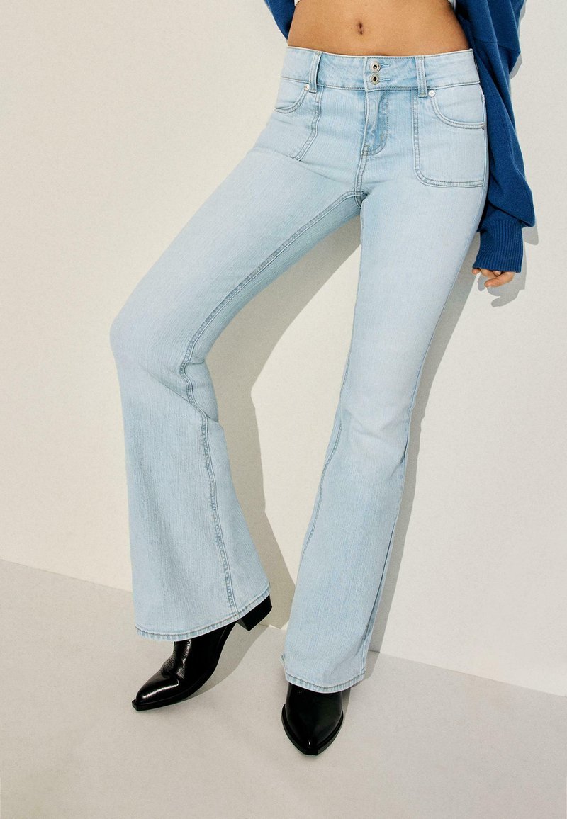 Jean évasé bleu clair avec fermeture à double bouton, porté avec des bottines noires à bouts pointus et un pull court bleu.