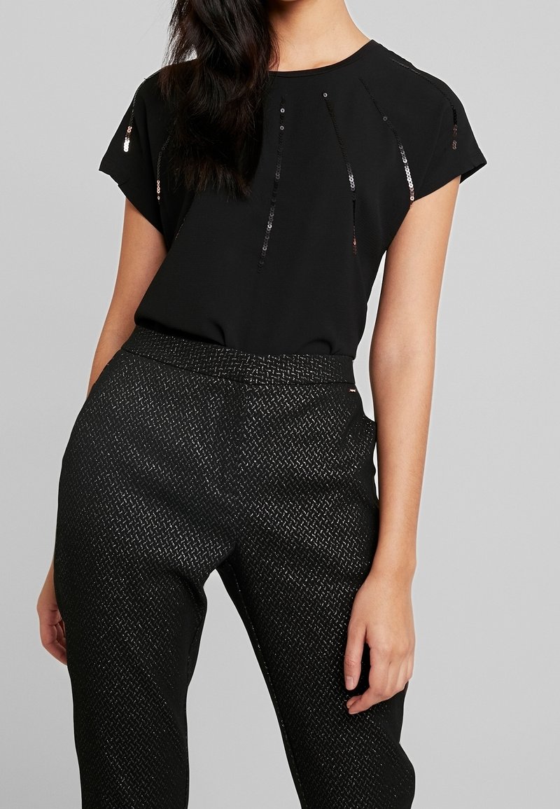 Femme portant un haut noir à manches courtes avec des sequins verticaux et un pantalon noir texturé taille haute, debout sur un fond gris.