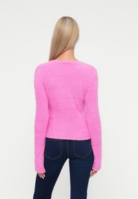 Puszysty różowy sweter z długimi rękawami i szerokim dekoltem, o miękkiej fakturze. W zestawieniu z ciemnoniebieskimi dżinsami. Czyste, minimalistyczne tło.