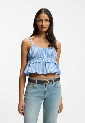 Vrouw met een lichtblauw gestreept, geruffleerd, mouwloos cropped topje en lichtblauwe jeans met een bruine riem, staand tegen een witte achtergrond.