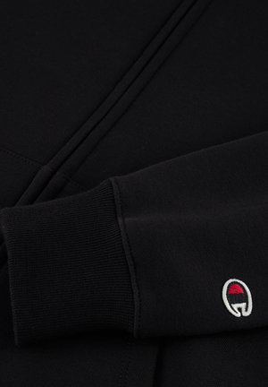 Sweatshirt noir avec fermeture éclair, poignets côtelés et un petit logo brodé en rouge, blanc et noir sur la manche. Texture de tissu douce.