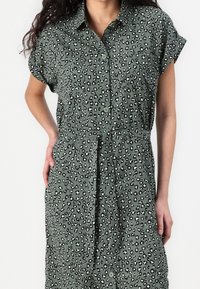 Femme portant une robe verte boutonnée avec un imprimé léopard noir et blanc et une ceinture nouée à la taille, debout avec une main sur la hanche.