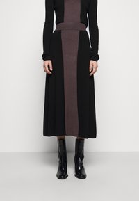 Nainen, joka on pukeutunut mustaan ja tummanliilaan pitkähihaiseen neulemekkoon, jossa on keskipaneeli, yhdistettynä kiiltäviin mustiin nilkkureihin, seisoo tasaisella lattialla.