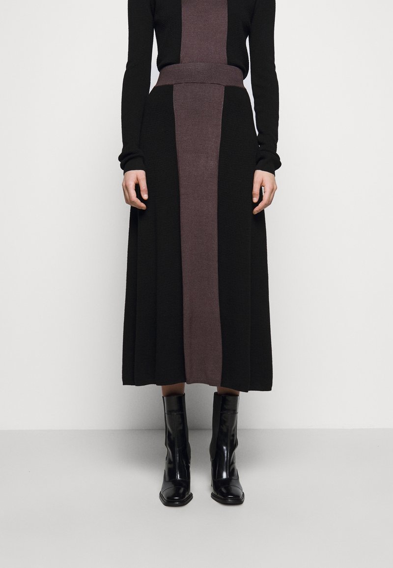 Nainen, joka on pukeutunut mustaan ja tummanliilaan pitkähihaiseen neulemekkoon, jossa on keskipaneeli, yhdistettynä kiiltäviin mustiin nilkkureihin, seisoo tasaisella lattialla.