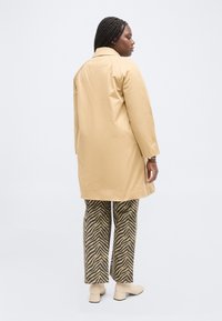 Γυναίκα με πλεξούδες, ντυμένη με μπεζ παλτό μήκους μέχρι το γόνατο, παντελόνι με ρίγες ζέβρας και μπεζ μποτάκια με τακούνι, που στέκεται μπροστά σε απλό λευκό φόντο.