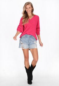 Femeie zâmbitoare cu păr lung, îmbrăcată într-un pulover roz cu decolteu în V, pantaloni scurți din jeans rupti, cizme negre până la jumătatea gambei și cercei roz în formă de diamant, mergând înainte.