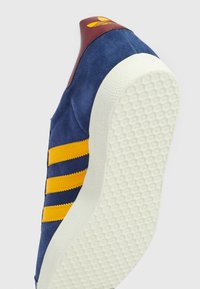 Blå mockasinsneaker med gula ränder, vinröd häl och texturerad vit gummisula. Sidvy visar tydlig form och konstruktion.