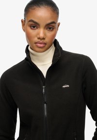 Zwarte fleece jas met rits en een hoge kraag, met een logo op de borst en een gladde, zachte textuur. Ideaal om als tussenlaag te dragen.