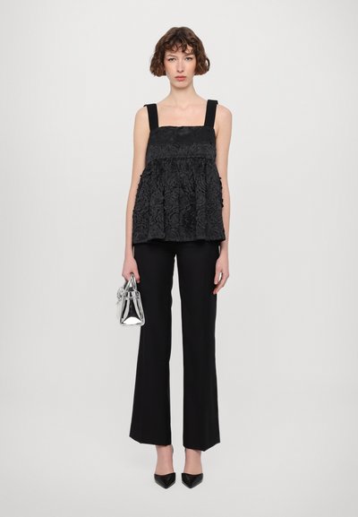 Top din dantelă florală neagră cu bretele late, asociat cu pantaloni negri cu talie înaltă. Modelul ține o geantă mică din metal.