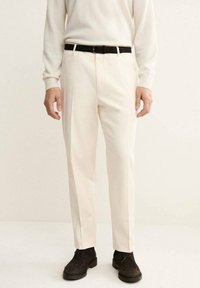 Pantalones beige de corte recto, con un cinturón negro. Tejido texturizado y diseño de frente plano. Llevados con zapatos oscuros.