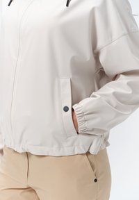 Lys beige cropped jakke med elastiske manchetter, lynlåslukning og to sidelommer. Fremstillet af glat, vandafvisende materiale.