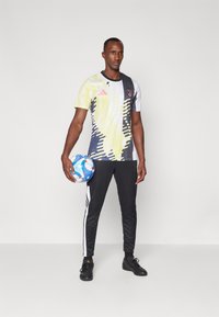 T-shirt de sport à motifs graphiques jaunes et noirs avec des manches courtes, assorti à un pantalon noir avec des rayures blanches, et des chaussures de football noires.