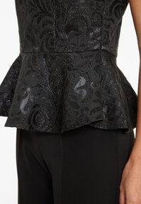 Gros plan sur un haut peplum noir avec un motif floral en brocart brillant, associé à un pantalon noir ajusté uni.