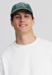 Felpa bianca con collo rotondo. Un cappellino verde presenta la scritta "NEW JERSEY" e "EAST COAST" in lettere bianche. Design semplice, vestibilità rilassata.