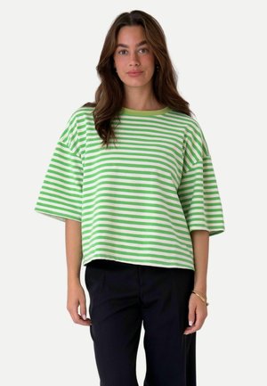 Mujer con cabello castaño largo, vistiendo una camisa holgada de manga corta a rayas verdes y blancas y pantalones negros, de pie frente a un fondo liso.