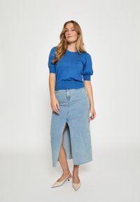 Zila adīta īsām piedurknēm blūze, apvienota ar gaišu džinsa garo svārku ar priekšējo šķēlumu. Modelis valkā beigu krāsas ar norādītu pirkstu augstpapēžu kurpes.