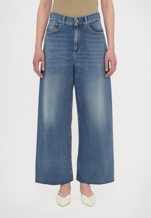 Persoon draagt een hoge taille, wijd uitlopende blauwe spijkerbroek met vervaagde voorkant en ruwe zomen, gecombineerd met een beige top en witte pumps met een puntige neus.