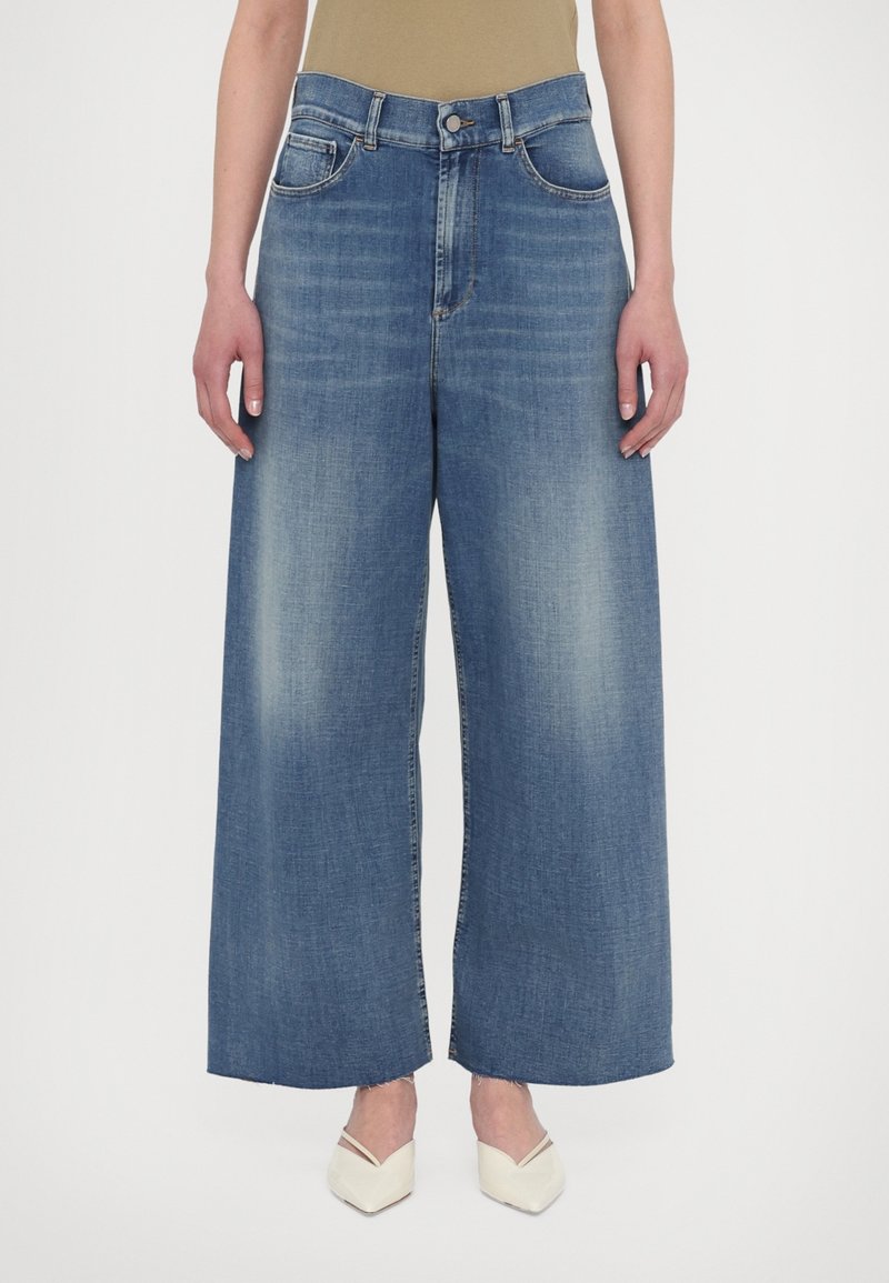 Personne portant un jean taille haute à jambes larges bleu avec un effet délavé à l'avant et des ourlets brut, associé à un haut beige et des talons pointus blancs.