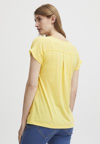 Gul kortærmet bluse med rund halsudskæring, plisseret detalje på ryggen, og en glat, let tekstur. Sammen med denim jeans.