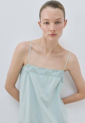 Mlajša ženska s svetlo poltjo in modrimi očmi nosi svetlomodro zgornji del brez rokavov z čipkastim robom, stoji ob enostavni svetli podlagi.