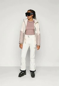 Σακάκι σκι σε κρέμα και ανοιχτό γκρι, υφασμάτινη μπλούζα από κάτω, άσπρο παντελόνι και μαύρα μποτάκια σκι. Αξεσουάρ περιλαμβάνουν σκούρα γυαλιά σκι.