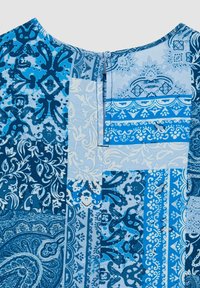 Blusa in patchwork blu con vari motivi intricati, scollatura rotonda e chiusura con bottone nel retro. Miscela di tonalità di blu chiaro e scuro.