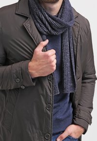 Tmavá péřová bunda s knoflíkovým zapínáním, doplněná žebrovanou námořnicky modrou šálou. Pod ní je přiléhavý modrý svetr. Je patrná texturovaná tkanina.