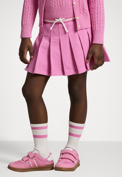 Rosa gestrickte Strickjacke, passende rosa plissierte Rock mit elastischem Bund und rosa Sneakers mit weißen Akzenten und Klettverschlüssen. Gestreifte Socken.