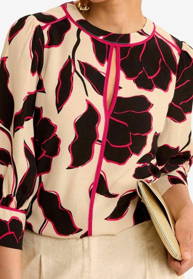 Blouse beige avec motif abstrait de feuilles noires et roses, encolure en trou de serrure, manches bouffantes, associée à un pantalon beige et une pochette dorée.