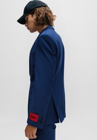 Blaues Sakko mit einem einzelnen Knopfverschluss, schmalen Revers und einem roten Etikett mit der Aufschrift "HUGO" am Ärmel. Das Modell trägt eine schwarze Mütze.