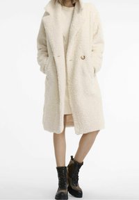 Kremowy, puszysty płaszcz z długim włosem, z bocznymi kieszeniami, dużym kołnierzem i pojedynczym zapięciem na guzik. Noszony na prążkowanej sukience i masywnych, sznurowanych butach.