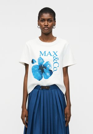 Nő fehér pólóban, amin kék virág és a „MAX & Co.” felirat látható, magas derekú, pliszírozott kék szoknyával és karika fülbevalóval.