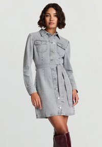 Vestito camicia in denim grigio con maniche lunghe, davanti con bottoni, collo appuntito, due tasche sul petto e cintura abbinata. Texture liscia, vestibilità strutturata.